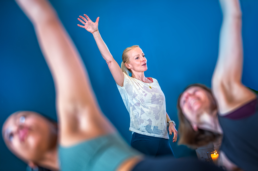 Foto einer Yogalehrerin, die Yogaübende mit zeitgemäßem Yogaunterricht anleitet - Führung durch Klarheit: Bewusste Gestik unterstützt die Anleitung und gibt der Gruppe Sicherheit. Zeitgemäßer Yogaunterricht bedeutet, den Raum zu halten – mit Präsenz, nicht mit Performance.